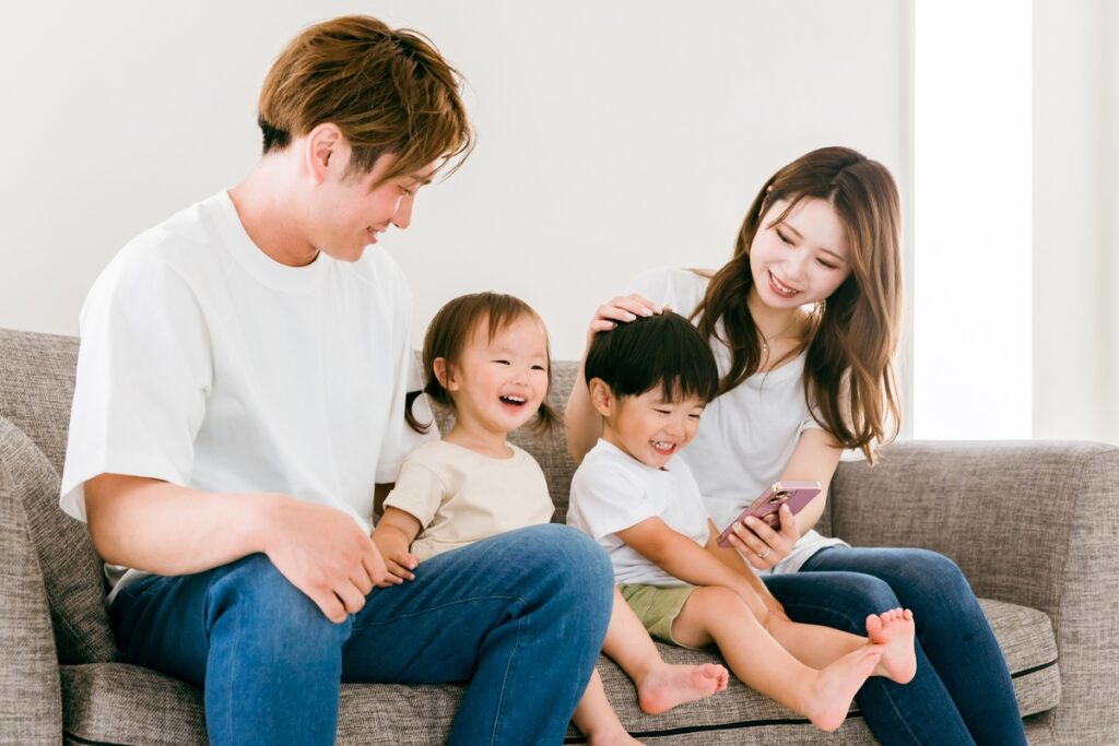 リビングのソファで子供たちとスマートフォンを見ながら笑顔で過ごす、白Tシャツとデニムの若い夫婦
