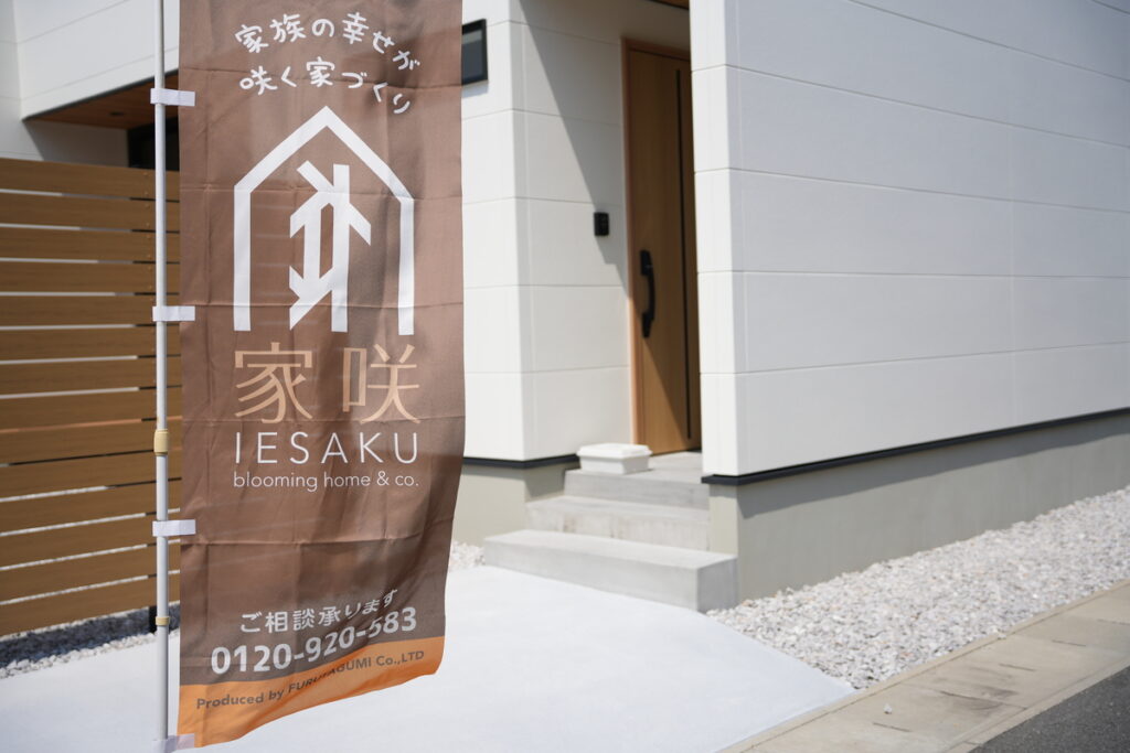 「家族の幸せが咲く家づくり 家咲 IESAKU」と書かれたブラウンののぼり旗が立つ白い外壁の新築住宅