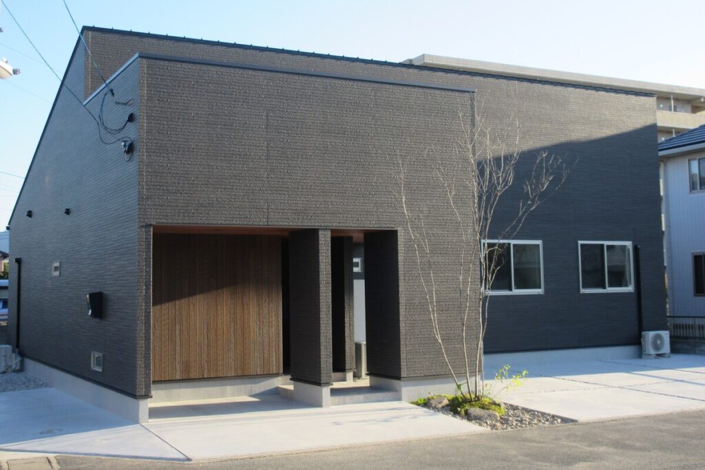 シンプルな箱型デザインの注文住宅の外観。ダークグレーの外壁と木目の玄関が印象的な住まい。