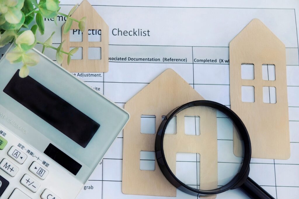 住宅のチェックリストの上に置かれた、家を模した木製オブジェと虫眼鏡、電卓。