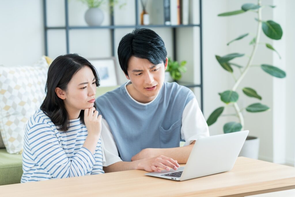 ノートパソコンの画面を深刻な表情で覗き込み、建築現場の状況を相談する若い夫婦