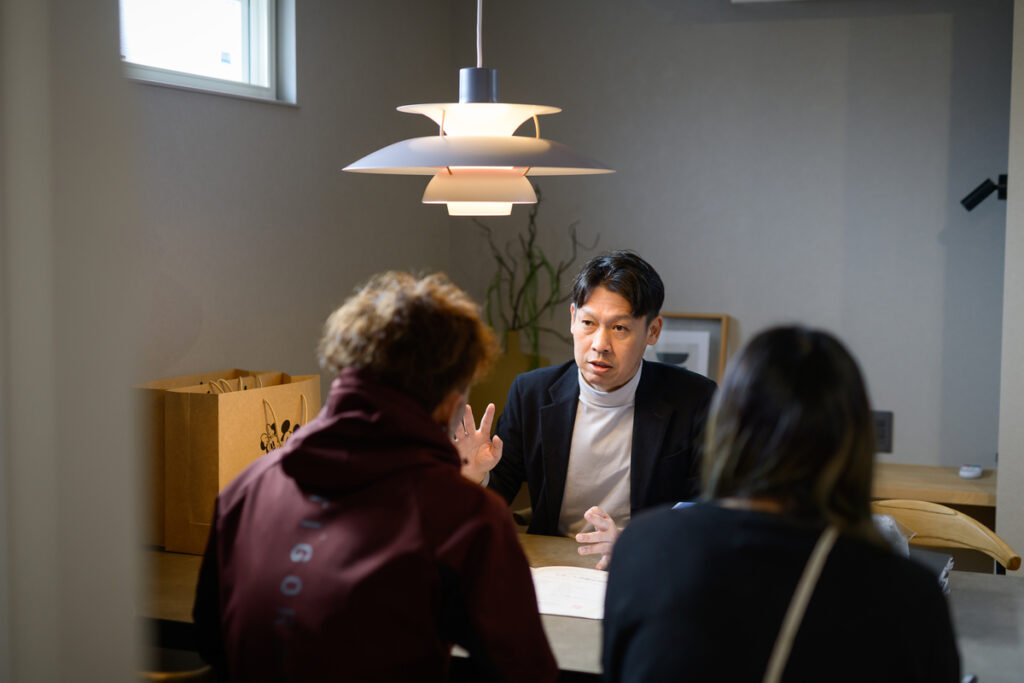 落ち着いた照明のリビングで、住宅ローンの相談に乗る専門スタッフと夫婦の打ち合わせ風景。