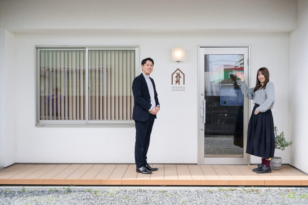 清潔感のある白い外壁とウッドデッキが特徴的な「IESAKU SHOWROOM」の入り口で、来場者を笑顔で出迎えるスタッフ。